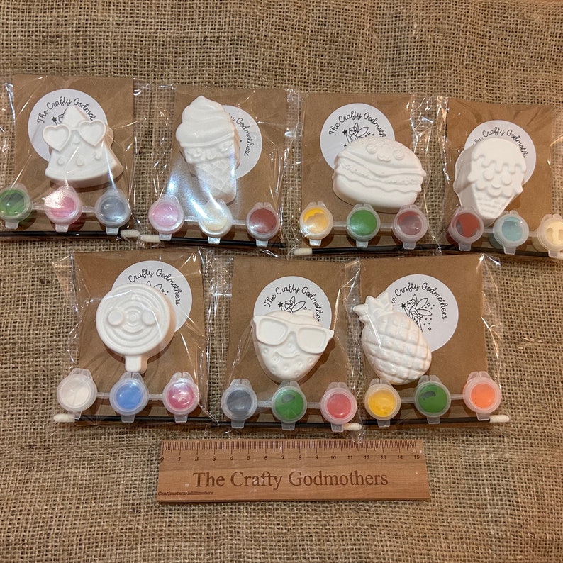 May include: Eight paint-your-own ceramic ornaments in individual clear plastic bags with paint pots and a paintbrush. The ornaments are shaped like a pineapple, ice cream cone, sunglasses, a slice of watermelon, a lollipop, a cake, a flower, and a pair of hearts. The bags are labeled "The Crafty Godmothers".