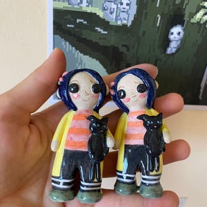 May include: Two ceramic figurines of a girl with blue hair, wearing a yellow raincoat, orange and black striped shirt, black pants, and white socks. Each figurine is holding a black cat.