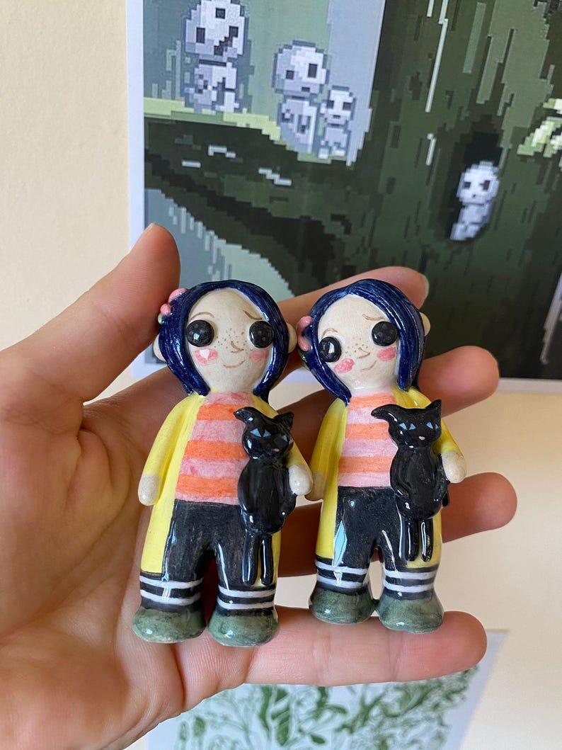 May include: Two ceramic figurines of a girl with blue hair, wearing a yellow raincoat, orange and black striped shirt, black pants, and white socks. Each figurine is holding a black cat.