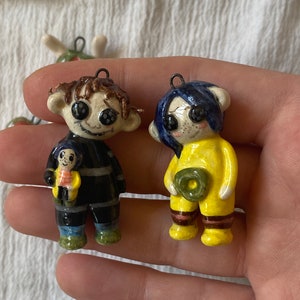 May include: Two ceramic figurines with button eyes. One figure is in a black outfit and holding a smaller figure. The other figure is in a yellow outfit and holding a green object. Both have a loop for hanging.