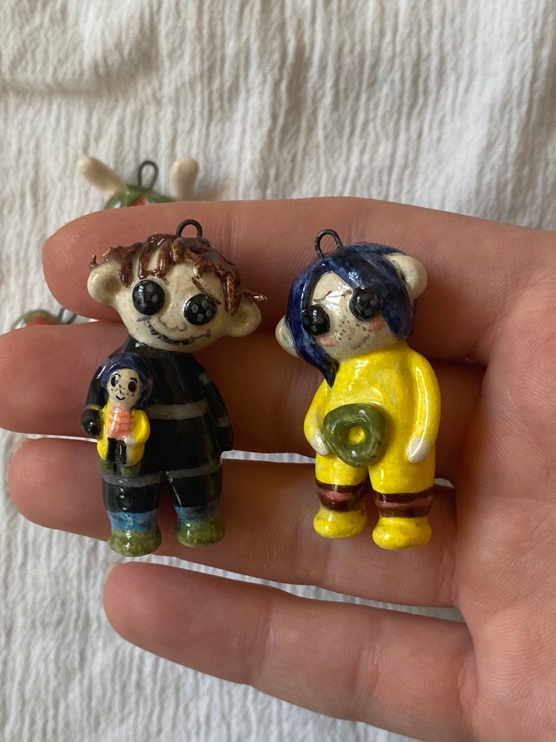 May include: Two ceramic figurines with button eyes. One figure is in a black outfit and holding a smaller figure. The other figure is in a yellow outfit and holding a green object. Both have a loop for hanging.