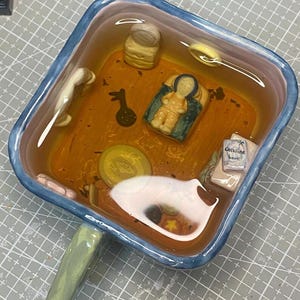 May include: A square ceramic dish with a blue rim and green handle, filled with a clear liquid and miniature figurines. The figurines include a baby, a book labeled "Coraline book", and other small objects. The dish has an orange and brown interior.