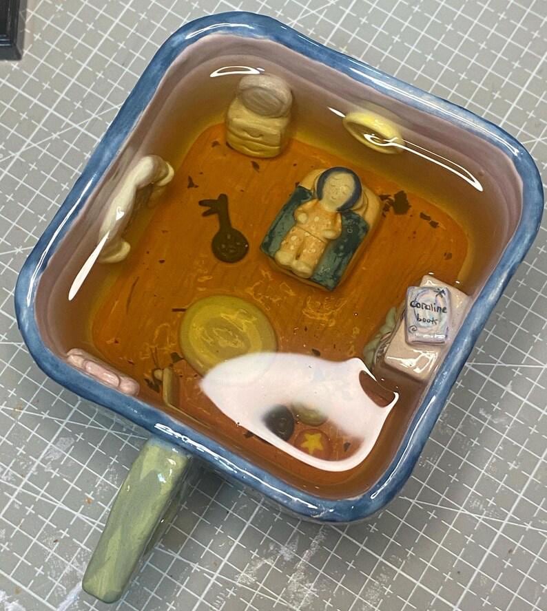 May include: A square ceramic dish with a blue rim and green handle, filled with a clear liquid and miniature figurines. The figurines include a baby, a book labeled "Coraline book", and other small objects. The dish has an orange and brown interior.