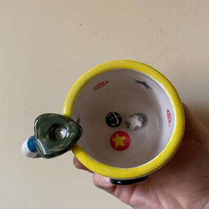 May include: A ceramic mug with a bright yellow rim and a dark green, triangular-shaped handle. The interior features a white base decorated with a black gear shift pattern, a red circle with a yellow star, and small red spiral and blue star motifs.