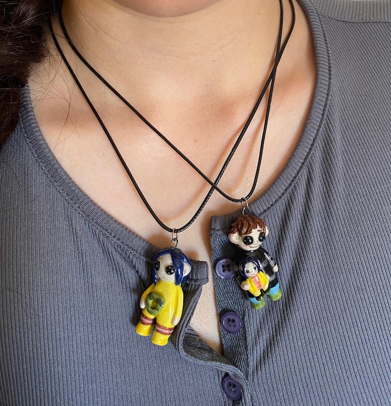 May include: Two black cord necklaces each feature a handmade doll pendant. One doll has blue hair and a yellow outfit, the other has brown hair and a black outfit, each holding a smaller doll. The pendants are attached to the cords.