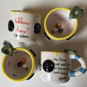 May include: Two white ceramic mugs with yellow rims and black button accents. One mug features the text "Welcome home! Coraline" and the other "Hey Jonesy Look what I found in Gramma's trunk Look familiar? Wybie". Both mugs contain small sculpted items.