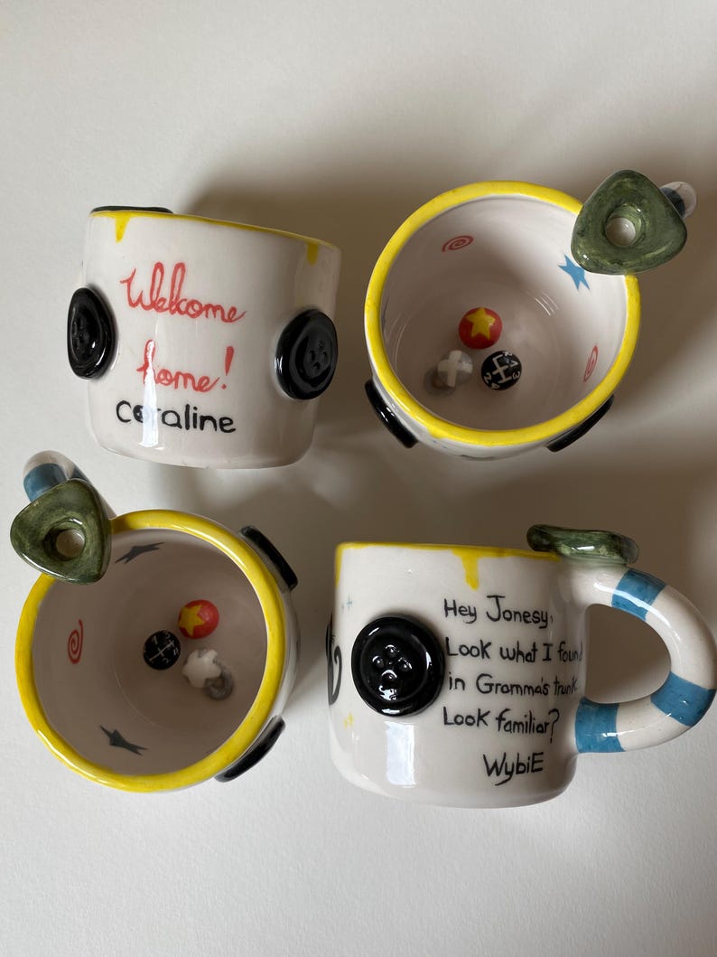 May include: Two white ceramic mugs with yellow rims and black button accents. One mug features the text "Welcome home! Coraline" and the other "Hey Jonesy Look what I found in Gramma's trunk Look familiar? Wybie". Both mugs contain small sculpted items.