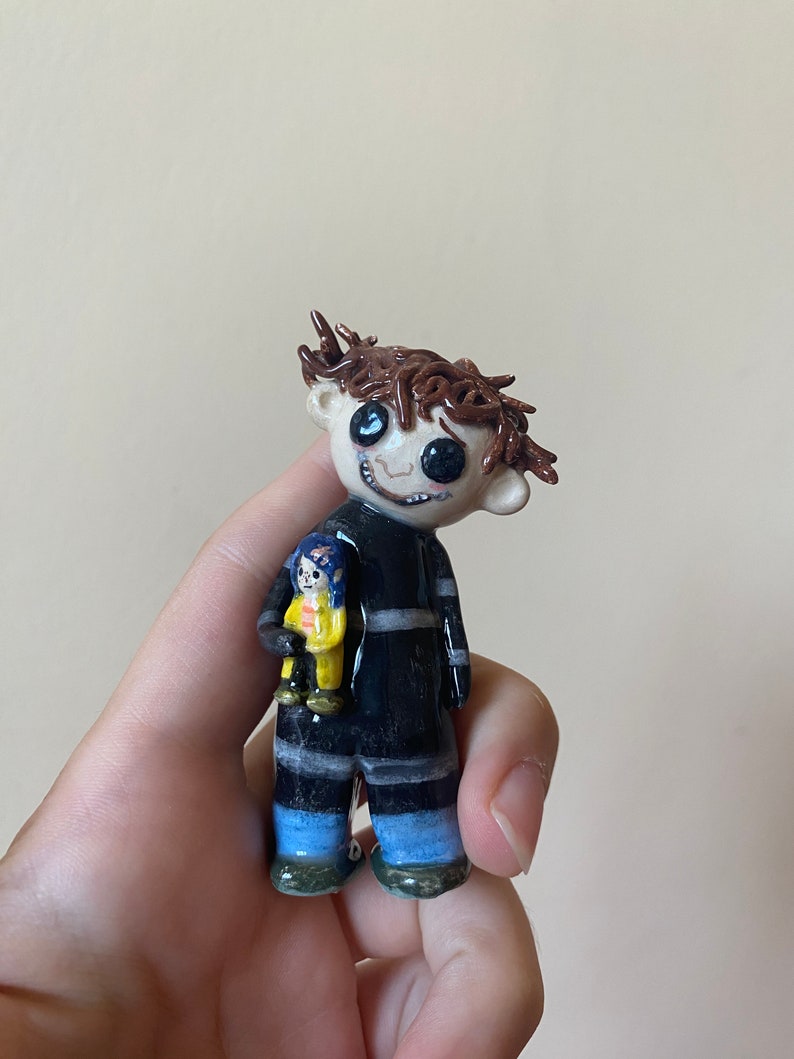 May include: A hand-painted ceramic figurine of a boy with brown hair, wearing a black and white striped shirt and blue pants. He is holding a smaller figurine of a girl in a yellow raincoat.