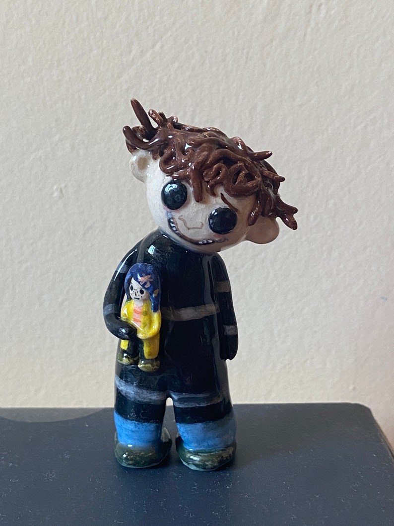 May include: A ceramic figurine of a person with brown hair, wearing a black and white striped shirt and blue pants, holding a smaller ceramic figurine of a person with blue hair and a yellow dress.