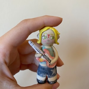 May include: A hand-painted ceramic figurine of a person with blonde hair, wearing a blue shirt and blue shorts with a white star, holding a pink and white electric guitar.
