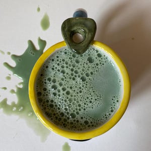 May include: A ceramic mug with a bright yellow rim and a dark green, triangular-shaped handle. The mug is filled with a frothy, light green liquid that has spilled onto the surface, creating a splatter pattern of green droplets around the base.