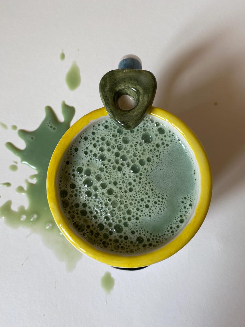 May include: A ceramic mug with a bright yellow rim and a dark green, triangular-shaped handle. The mug is filled with a frothy, light green liquid that has spilled onto the surface, creating a splatter pattern of green droplets around the base.
