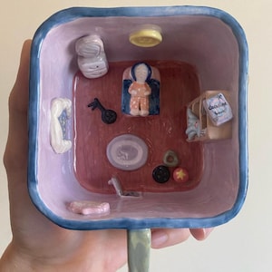May include: A whimsical ceramic mug with a square interior, painted in shades of pink, blue, and red. The interior features miniature ceramic objects, including a figure, a musical instrument, and a small box with the text "Coraline book".