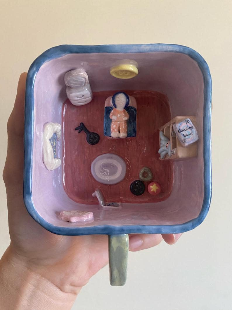 May include: A whimsical ceramic mug with a square interior, painted in shades of pink, blue, and red. The interior features miniature ceramic objects, including a figure, a musical instrument, and a small box with the text "Coraline book".