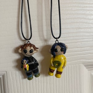 May include: Two handmade pendant necklaces featuring character figures. One figure is in a black and gray outfit, holding a smaller figure. The other figure is in a yellow outfit. Both pendants are attached to black cords.