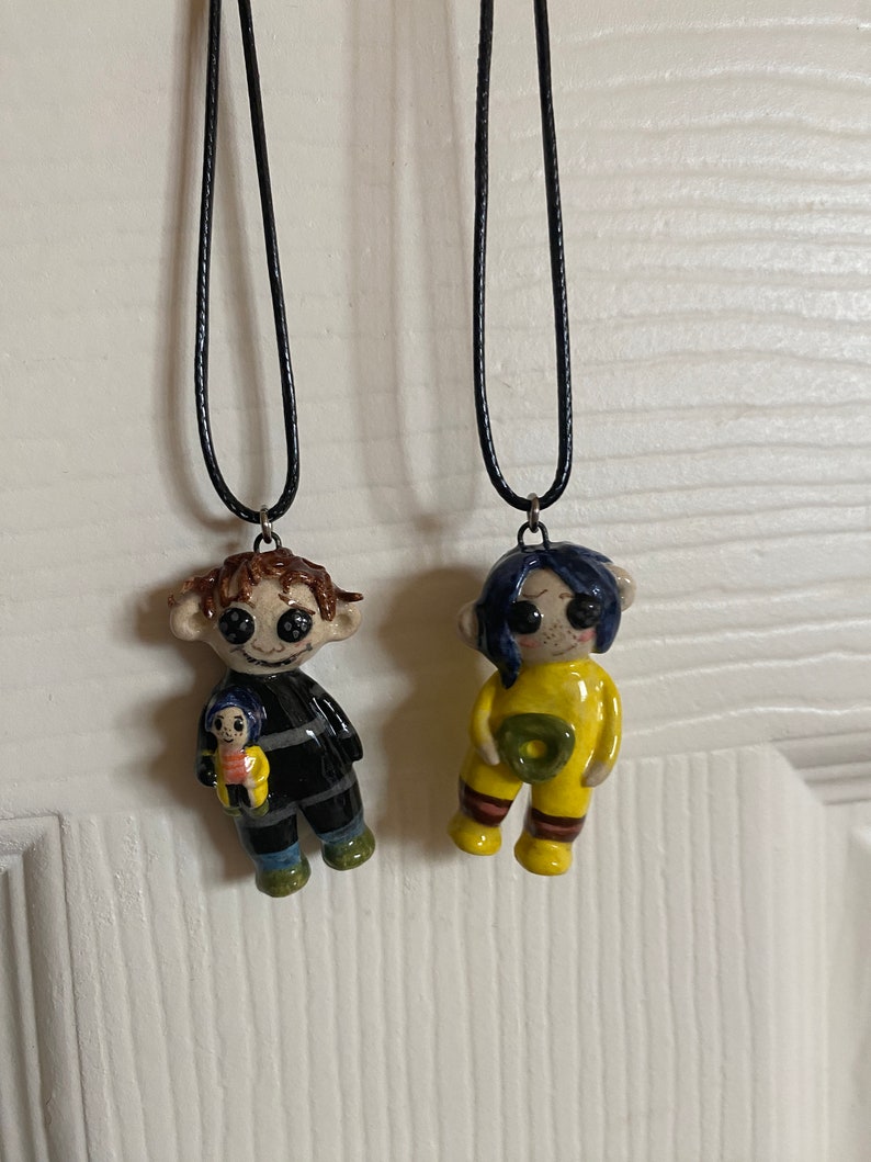 May include: Two handmade pendant necklaces featuring character figures. One figure is in a black and gray outfit, holding a smaller figure. The other figure is in a yellow outfit. Both pendants are attached to black cords.