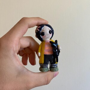 May include: A hand-painted ceramic figurine of a girl with black hair, wearing a yellow jacket, black pants, and green shoes. She is holding a black cat in her arms.