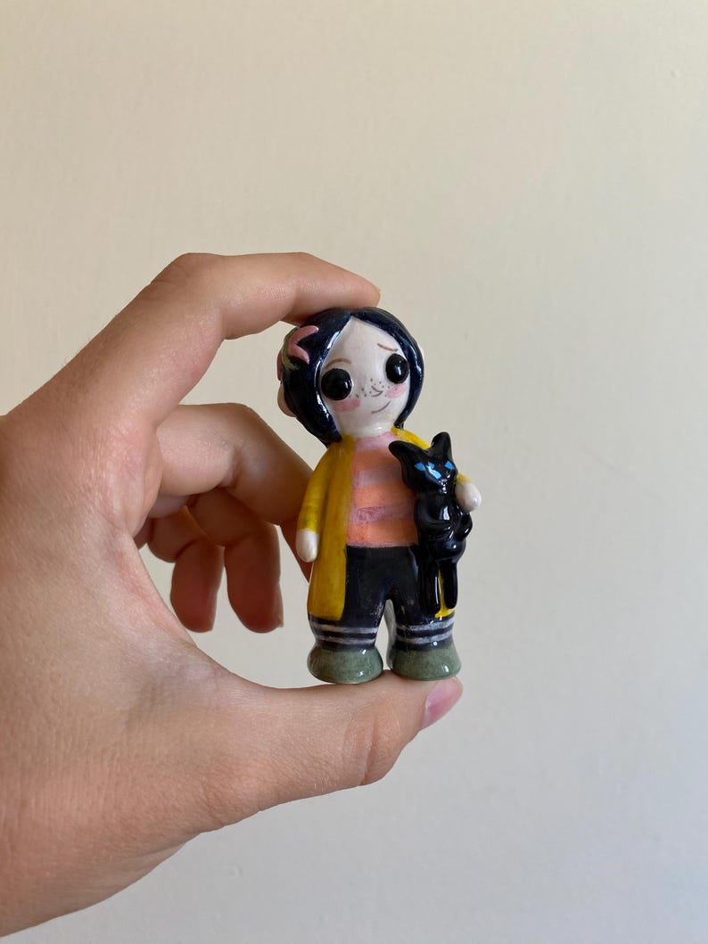 May include: A hand-painted ceramic figurine of a girl with black hair, wearing a yellow jacket, black pants, and green shoes. She is holding a black cat in her arms.