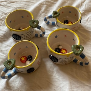May include: Three ceramic mugs with cream-colored interiors and exteriors, featuring yellow rims, blue and white striped handles, and green button-shaped accents. The mugs display black text reading Hey Jonesy, Look what I found in Gramma's trunk. Look familiar? Wybie.