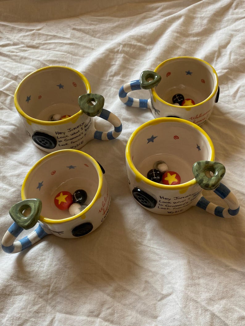 May include: Three ceramic mugs with cream-colored interiors and exteriors, featuring yellow rims, blue and white striped handles, and green button-shaped accents. The mugs display black text reading Hey Jonesy, Look what I found in Gramma's trunk. Look familiar? Wybie.
