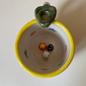 May include: A ceramic mug with a bright yellow rim and a white interior featuring blue star and red swirl patterns. Inside the mug are three small decorative spheres: one red with a yellow star, one black with a gear shift pattern, and one white star.