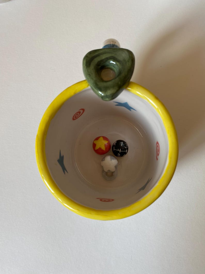 May include: A ceramic mug with a bright yellow rim and a white interior featuring blue star and red swirl patterns. Inside the mug are three small decorative spheres: one red with a yellow star, one black with a gear shift pattern, and one white star.