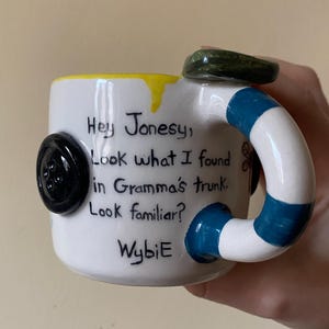 May include: A white ceramic mug featuring a black button attached to the side and a handle with blue and white stripes. The mug has yellow glaze at the rim and text that reads: Hey Jonesy, Look what I found in Gramma's trunk. Look familiar? WybiE.