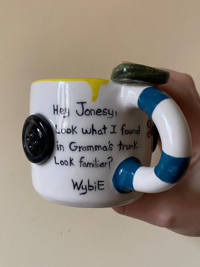 May include: A white ceramic mug featuring a black button attached to the side and a handle with blue and white stripes. The mug has yellow glaze at the rim and text that reads: Hey Jonesy, Look what I found in Gramma's trunk. Look familiar? WybiE.