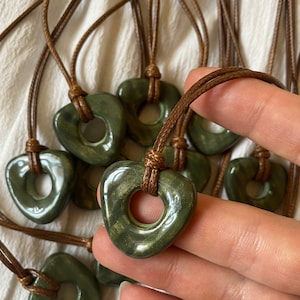 May include: A collection of green heart-shaped pendants with a hole in the center, strung on brown cords. The pendants have a polished, glossy finish. The cords are knotted at the top. The background is a white fabric.