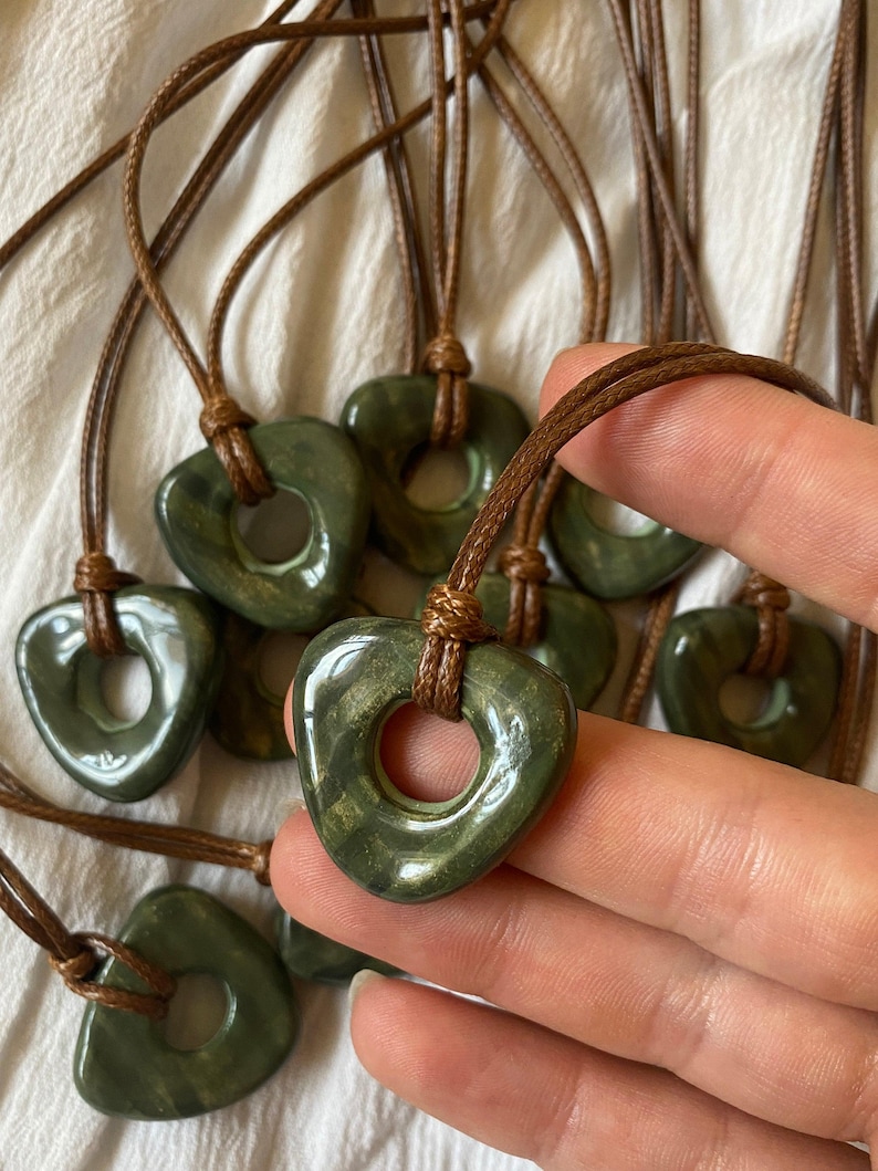 May include: A collection of green heart-shaped pendants with a hole in the center, strung on brown cords. The pendants have a polished, glossy finish. The cords are knotted at the top. The background is a white fabric.