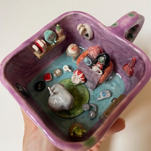 May include: A handmade ceramic dish with a purple exterior and a detailed interior scene. The interior features miniature sculptures of figures, objects, and a pond. The dish has a handle with green polka dots.