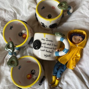May include: Three white ceramic mugs with yellow rims and blue and white striped handles. Each mug features black button accents, small painted stars, and swirls. One mug displays the text: Hey Jonesy, Look what I found in Gramma's trunk. Look familiar? WybiE.