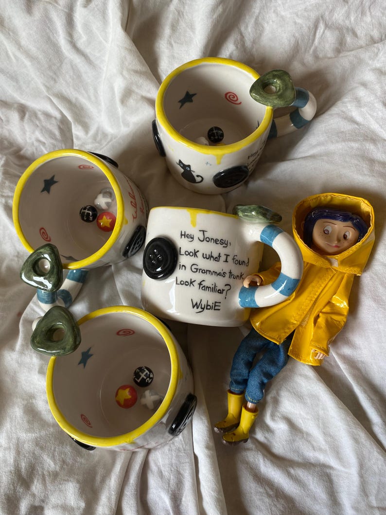May include: Three white ceramic mugs with yellow rims and blue and white striped handles. Each mug features black button accents, small painted stars, and swirls. One mug displays the text: Hey Jonesy, Look what I found in Gramma's trunk. Look familiar? WybiE.