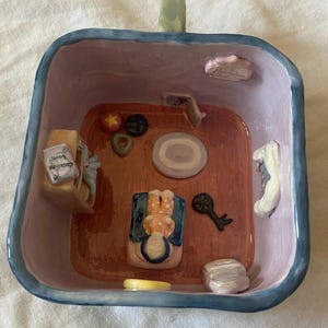 May include: A whimsical, hand-painted ceramic bowl with a miniature room scene inside. The interior features a tiny figure, furniture, and decorative elements. The bowl has a blue rim and a green handle.