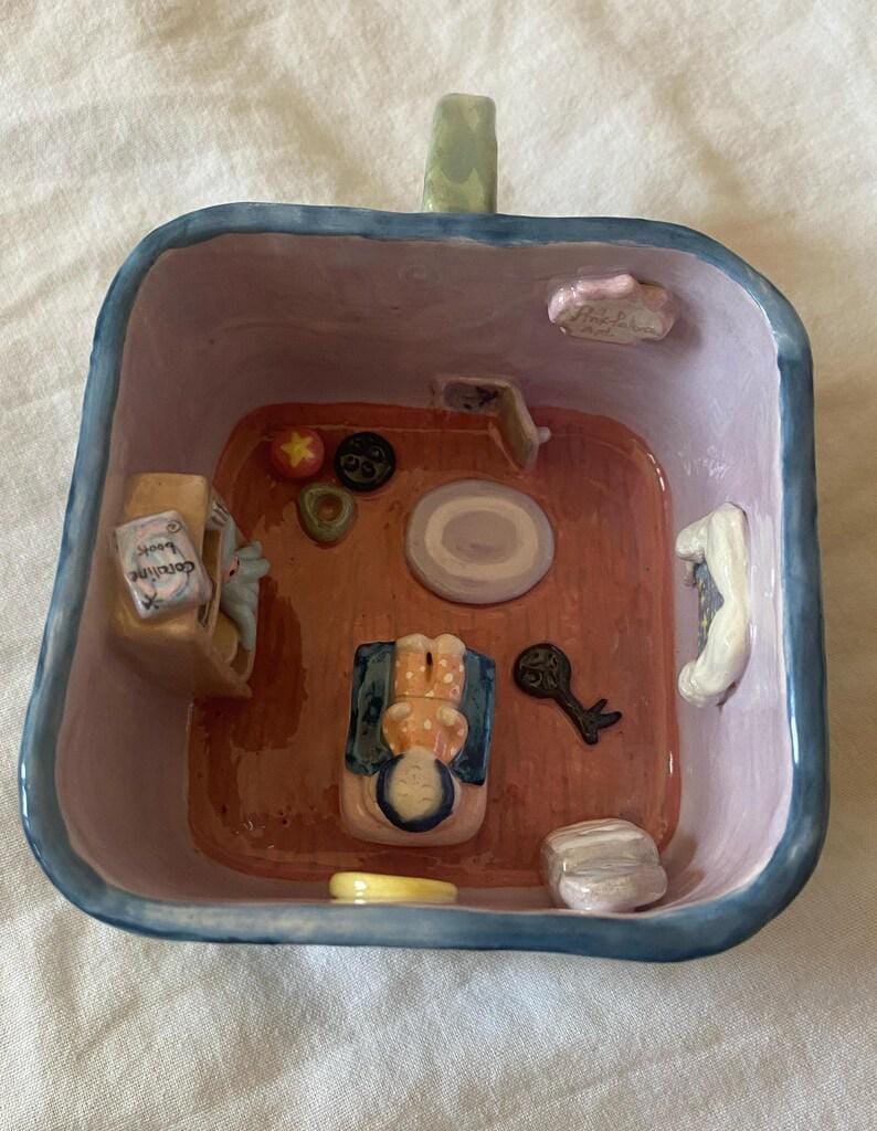 May include: A whimsical, hand-painted ceramic bowl with a miniature room scene inside. The interior features a tiny figure, furniture, and decorative elements. The bowl has a blue rim and a green handle.