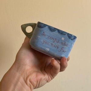 May include: A hand-painted ceramic mug with a light blue exterior and a green handle. The mug features the text "Be careful what you wish for" in handwritten style, along with decorative star and wave accents. The mug is held by a person.