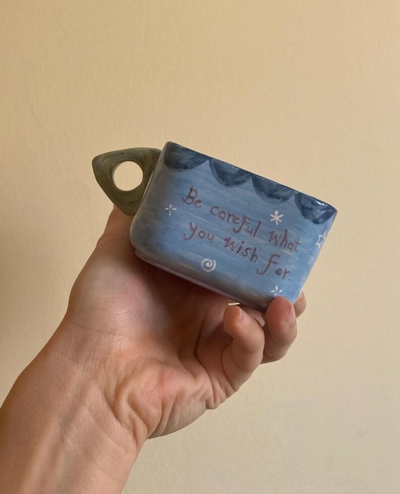 May include: A hand-painted ceramic mug with a light blue exterior and a green handle. The mug features the text "Be careful what you wish for" in handwritten style, along with decorative star and wave accents. The mug is held by a person.