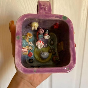 May include: A square ceramic mug with a purple glazed exterior featuring a star embossed on the handle. The interior is a miniature diorama containing small sculpted figures, including two people, a peppermint candy, and various tiny objects.