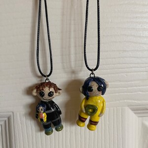 May include: Two handmade doll pendant necklaces. One doll wears a black and gray striped outfit and holds a smaller doll. The other doll wears a yellow jumpsuit with a green circle. Both dolls have button eyes and are suspended from black cords.