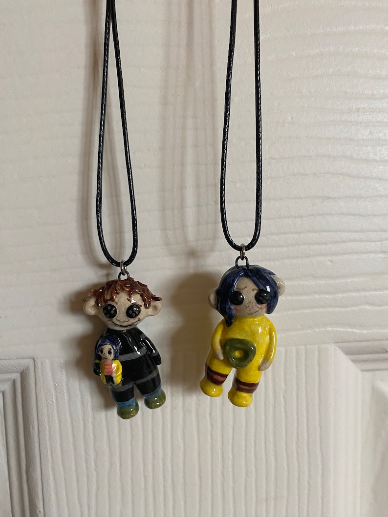 May include: Two handmade doll pendant necklaces. One doll wears a black and gray striped outfit and holds a smaller doll. The other doll wears a yellow jumpsuit with a green circle. Both dolls have button eyes and are suspended from black cords.