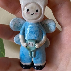 May include: A hand-painted ceramic figurine of a blue fairy with wings, holding a small green creature. The fairy has a white hat and a smiling face.