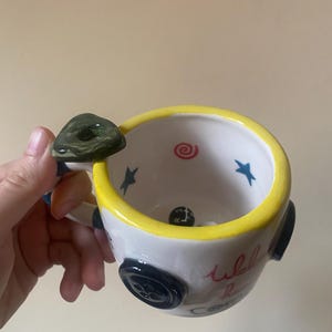 May include: A white ceramic mug with a yellow rim, featuring a dark green button-shaped handle. The exterior is decorated with black buttons and red cursive text. Inside, the mug displays blue stars, a red spiral, and a black gear shift knob.