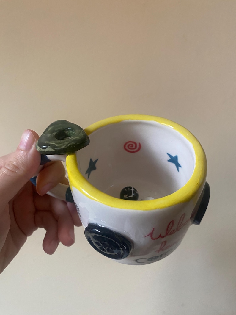 May include: A white ceramic mug with a yellow rim, featuring a dark green button-shaped handle. The exterior is decorated with black buttons and red cursive text. Inside, the mug displays blue stars, a red spiral, and a black gear shift knob.