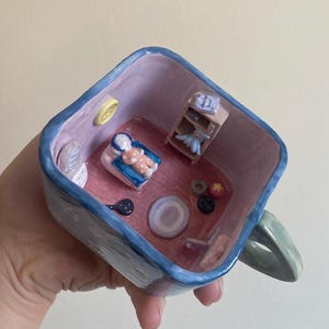 May include: A whimsical ceramic mug with a miniature room scene inside. The interior features a bed with a doll, a small shelf with a book and a toy, and other tiny decorative elements. The mug has a blue rim and handle.