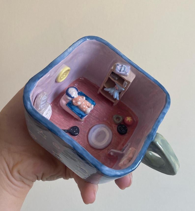 May include: A whimsical ceramic mug with a miniature room scene inside. The interior features a bed with a doll, a small shelf with a book and a toy, and other tiny decorative elements. The mug has a blue rim and handle.