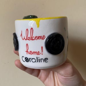 May include: A white ceramic mug featuring a yellow drip design along the top rim. The mug is decorated with black button appliques on the sides and the red text "Welcome home!" above the black text "Coraline" with a button icon for the letter O.