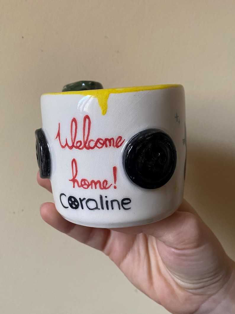 May include: A white ceramic mug featuring a yellow drip design along the top rim. The mug is decorated with black button appliques on the sides and the red text "Welcome home!" above the black text "Coraline" with a button icon for the letter O.