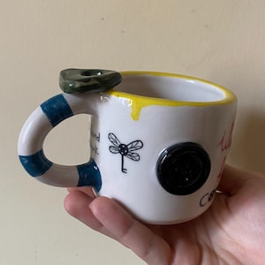 May include: A white ceramic mug with a yellow rim, a blue and white striped handle, and a dark green stone accent on top. The mug features a black dragonfly illustration, a black button attached to the side, and black text that reads I found trunk.