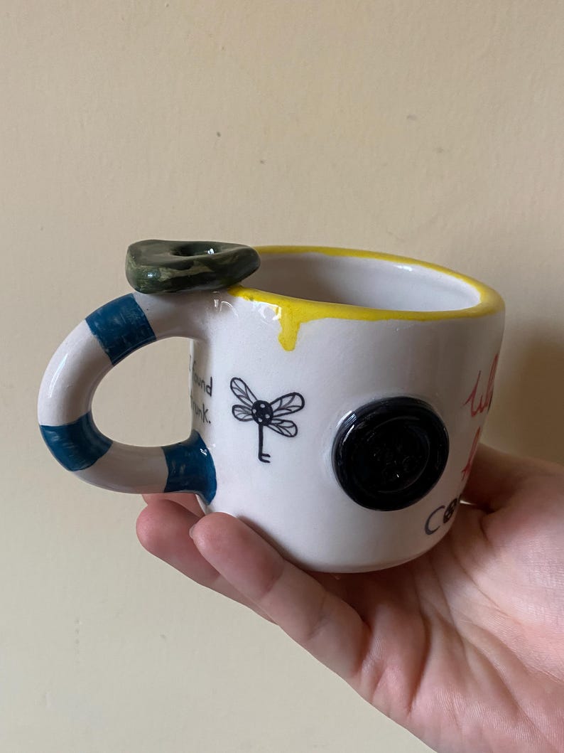 May include: A white ceramic mug with a yellow rim, a blue and white striped handle, and a dark green stone accent on top. The mug features a black dragonfly illustration, a black button attached to the side, and black text that reads I found trunk.
