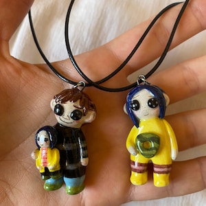 May include: Two handmade pendant necklaces featuring cartoon character figures. One figure has brown hair and a black and gray striped shirt, holding a smaller figure. The other figure has blue hair and a yellow outfit. Both pendants are on black cords.